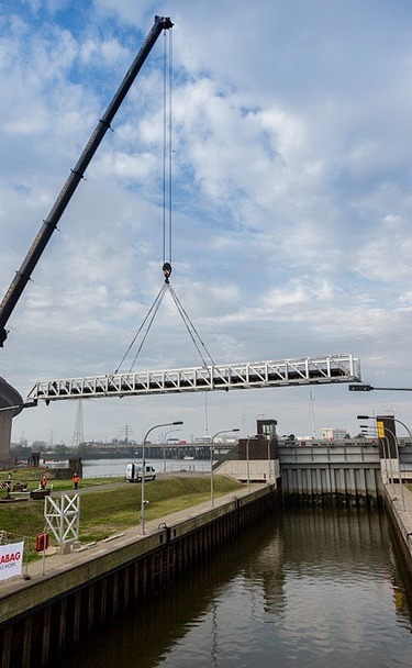 A mobile crane lifts a steel walkway over a lock at the Hamburg Harbor. The Köhlbrand Bridge and red container cranes are visible in the background, with STRABAG construction banners in the foreground.