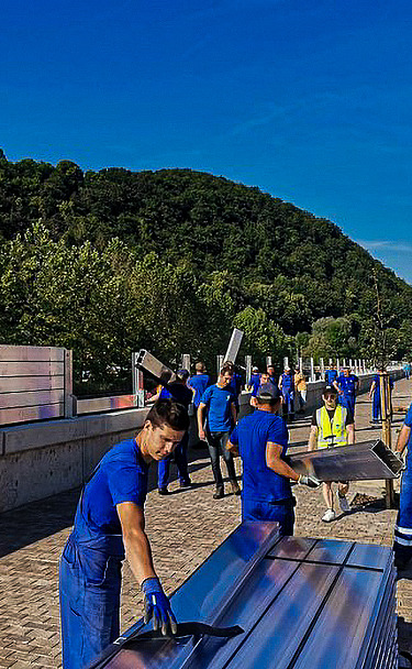 Gruppe von Arbeitern in blauen Uniformen, die große Metallplatten entlang einer Straßenabsperrung montieren. Die Szene umfasst einen gepflasterten Gehweg, Fahrzeuge auf der angrenzenden Straße und grüne Hügel im Hintergrund unter einem klaren blauen Himmel.