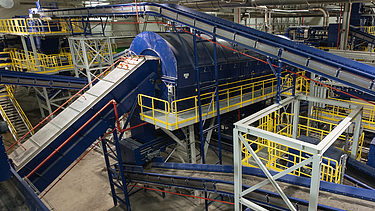 Aerial view of a large mechanical-biological waste treatment plant with green and orange buildings surrounded by landscape.