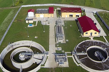 Aerial view of the Moinești wastewater treatment plant in Romania, showing circular clarifiers, treatment basins, control buildings with red roofs, and surrounding green fields within a fenced site.