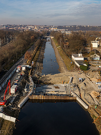Luftaufnahme eines im Bau befindlichen Flussschifffahrtskanals mit einem teilweise errichteten Schleusenbauwerk mit Stahlspundwänden, schweren Maschinen und Baumaterialien entlang des Ufers. In der Umgebung befinden sich eine parallel verlaufende Bahnlinie, Zufahrtsstraßen und vereinzelte Gebäude. Rechts ist ein breiter Fluss zu sehen und am Horizont eine entfernte Stadtlandschaft.