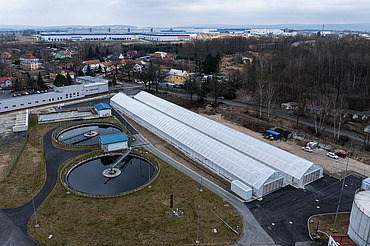 Aerial view of a wastewater treatment facility featuring two circular clarifiers and long greenhouse-like structures used for solar sludge drying, surrounded by roads, grass areas, and nearby residential and industrial buildings.