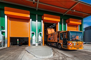 An orange refuse truck is shown at a modern recycling center with green and orange industrial bays. The vehicle is positioned at an open loading dock for waste disposal.