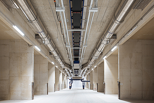 A long concrete corridor inside a facility with large silver metal pipes and a central conveyor system mounted to the ceiling.
