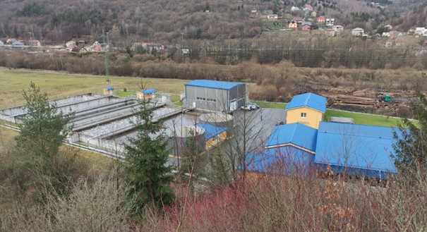 Aerial view of a water treatment facility with blue-roofed buildings and rectangular basins, surrounded by trees and hills in a rural area.