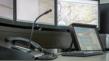 A dispatch workstation with multiple monitors showing maps and data, featuring a desktop microphone, a telephone handset, and a tablet controller.
