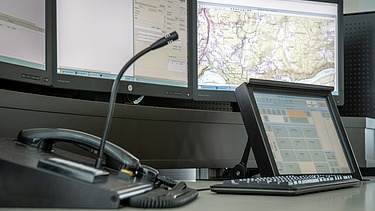 Ein Dispatcher-Arbeitsplatz mit mehreren Monitoren, auf denen Karten und Daten angezeigt werden, mit einem Tischmikrofon, einem Telefonhörer und einem Tablet-Controller.