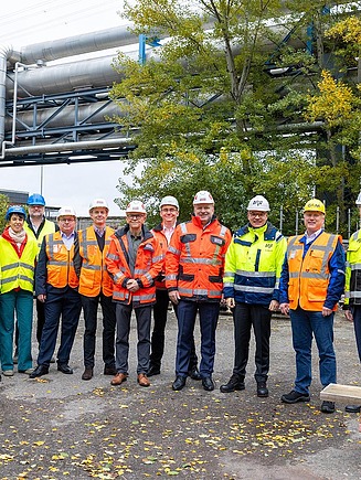Baustelle mit Betongebäuden, Stahlträgern und Bauarbeitern mit Sicherheitsausrüstung unter teils bewölktem Himmel