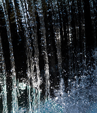Nahaufnahme eines senkrecht fallenden Wasserstrahls mit spritzenden und im Licht schimmernden Tropfen vor einem dunklen Hintergrund.