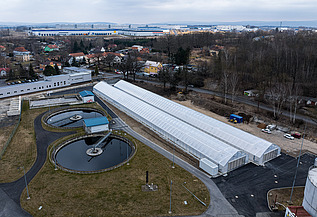 Aerial view of a wastewater treatment facility featuring two circular clarifiers and long greenhouse-like structures used for solar sludge drying, surrounded by roads, grass areas, and nearby residential and industrial buildings.