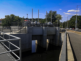 Concrete floodgate structure with three openings and metal railings, located next to a road. The structure includes vertical control rods and mechanical equipment on top. Trees and streetlights line the background under a clear blue sky.