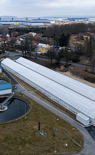 Aerial view of a wastewater treatment facility featuring two circular clarifiers and long greenhouse-like structures used for solar sludge drying, surrounded by roads, grass areas, and nearby residential and industrial buildings.
