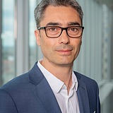 Person (Nedim) wearing a dark blue suit jacket over a white collared shirt, standing indoors in front of tall glass windows with a modern cityscape visible outside.