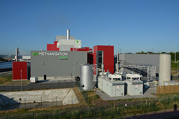 A detailed view focusing on the plant's technical installations, including a large silver tank, several containerized units, and an intricate pipe network. The main building with the 'BIOMÉTHANISATION' sign is visible in the background.