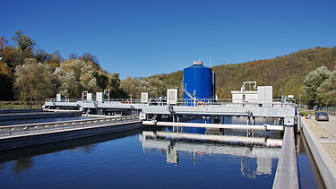 Concrete treatment basin filled with water at a wastewater treatment facility, with metal walkways, pipes, and a large blue cylindrical tank, surrounded by trees and hills under a clear sky.