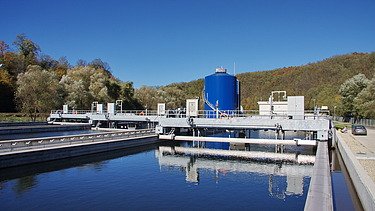 Concrete treatment basin filled with water at a wastewater treatment facility, with metal walkways, pipes, and a large blue cylindrical tank, surrounded by trees and hills under a clear sky.