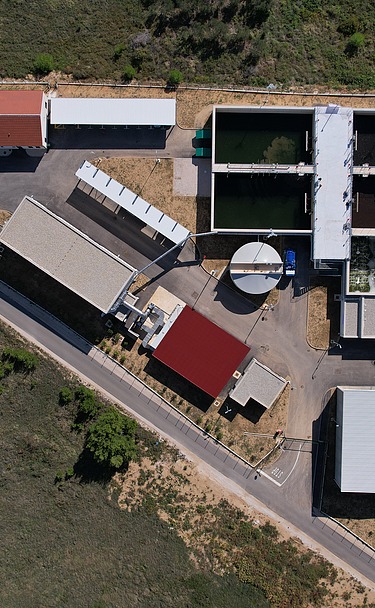 Top-down view of a wastewater treatment facility showing multiple rectangular treatment tanks, circular clarifier, and several buildings with red and white roofs, surrounded by green fields and access roads.