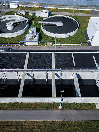 Aerial view of a modern wastewater treatment plant featuring rectangular sedimentation tanks in the foreground and two large circular clarifiers in the background, surrounded by green grass and facility buildings.