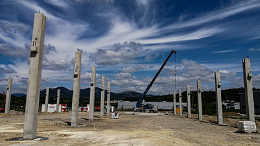 Baustelle mit mehreren aufrecht stehenden Betonsäulen, die in Reihen angeordnet sind und das Gerüst eines großen Bauwerks bilden. In der Mitte steht ein Kran, der Materialien anhebt. Der Hintergrund zeigt Hügel, verstreute Gebäude und einen teilweise bewölkten Himmel mit dramatischen Wolkenmustern.