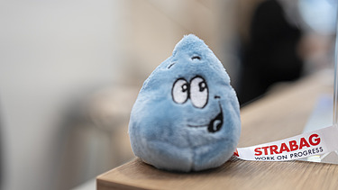 Close-up of a friendly, blue water droplet plush toy with a STRABAG ribbon, serving as a charming giveaway.
