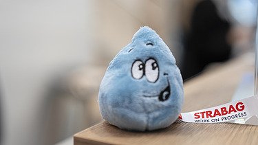 Nahaufnahme eines freundlichen, blauen Wassertropfen-Plüschtiers mit einem STRABAG-Band, das als charmantes Werbegeschenk dient.
