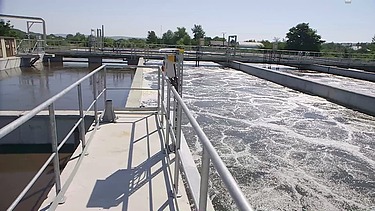 Blick entlang eines Weges in einer Kläranlage mit Belebungsbecken mit sprudelndem Wasser, Metallgeländern, Rohren und Kläranlagen bei klarem Tageslicht.