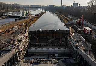 Baustelle einer Fahrrinnenschleuse an einem Fluss mit Stahlbetonkonstruktionen, Stahlspundwänden und Arbeitern. Im Hintergrund ist die Wasserstraße zu sehen, am Horizont sind städtische Gebäude zu erkennen.