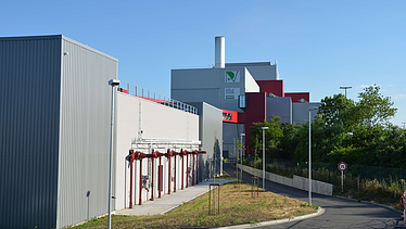 This image captures the rear of the facility, featuring a paved access road and a series of red pipes mounted on the gray building wall. A green 'Pole Énergie' logo can be seen higher up on the main building, and there is a speed limit sign by the roadside.