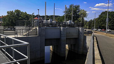 Schleusenbauwerk aus Beton mit drei Öffnungen und Metallgeländern, das an einer Straße liegt. Die Struktur umfasst vertikale Kontrollstangen und mechanische Geräte auf der Oberseite. Bäume und Straßenlaternen säumen den Hintergrund unter einem klaren blauen Himmel.