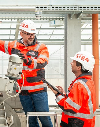 Two workers wearing orange and black safety jackets and white hard hats inside an industrial facility, inspecting and operating a mechanical component with one person holding a tablet and the other adjusting a motor.