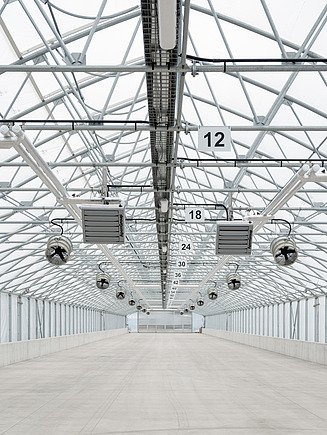 Longitudinal view inside a solar sludge drying hall featuring a ventilated greenhouse structure, roof-mounted ventilation equipment, circulation fans, and heating units. The hall provides controlled conditions for the solar drying of dewatered sewage sludge prior to its use as a renewable energy source.