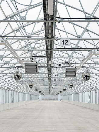 Längsansicht einer Solartrocknungshalle mit Gewächshauskonstruktion, Dachlüftung, Umluftventilatoren und Heizgeräten. Die technischen Einrichtungen schaffen optimale Bedingungen für die Trocknung von entwässertem Klärschlamm.