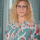 Person (Aniko), die eine gemusterte blaugrüne Bluse mit roten, weißen und blauen abstrakten Mustern trägt, steht in einem Innenraum vor hohen Glasfenstern, aus denen eine moderne Stadtlandschaft zu sehen ist.