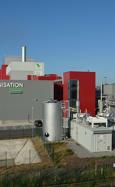 A detailed view focusing on the plant's technical installations, including a large silver tank, several containerized units, and an intricate pipe network. The main building with the 'BIOMÉTHANISATION' sign is visible in the background.