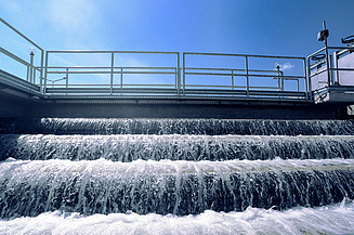 Multi-tiered water cascade at a treatment facility, with foaming water flowing over steps beneath a metal railing structure under a clear blue sky.