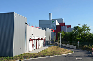 This image captures the rear of the facility, featuring a paved access road and a series of red pipes mounted on the gray building wall. A green 'Pole Énergie' logo can be seen higher up on the main building, and there is a speed limit sign by the roadside.