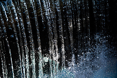 Nahaufnahme eines senkrecht fallenden Wasserstrahls mit spritzenden und im Licht schimmernden Tropfen vor einem dunklen Hintergrund.