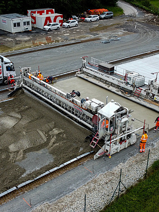 Construction site at a wastewater treatment facility showing workers and heavy machinery installing large concrete slabs for solar sludge drying units, with trucks and equipment in the background.
