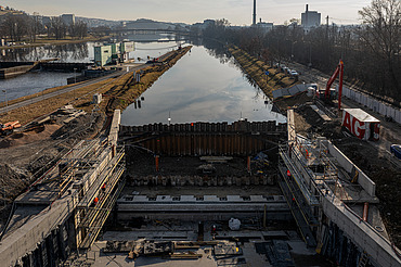 Construction site of a navigation channel lock along a river, showing reinforced concrete structures, steel sheet piling, and workers, with waterway extending into the background and urban buildings visible on the horizon.