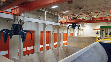An indoor industrial facility featuring large blue overhead claw cranes positioned over concrete waste pits and orange loading bay doors.