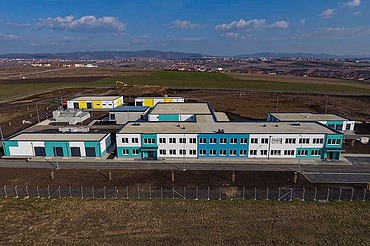 Aerial view of a modern water treatment plant in Prishtina, featuring multiple low-rise buildings with white and turquoise facades, flat roofs, and surrounding paved roads. The facility is located in a large open area with construction equipment visible in the background and distant hills under a partly cloudy sky.