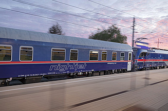 Blue night train speeding along railway tracks at dusk, with motion blur and overhead power lines.