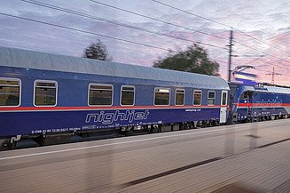 Blauer Nachtzug, der in der Dämmerung über die Bahngleise fährt, mit Bewegungsunschärfe und Überlandleitungen.
