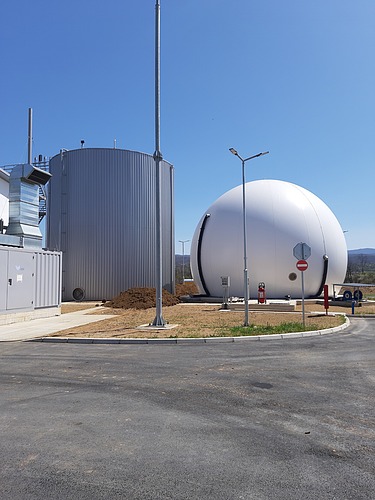 A close-up view of a wastewater treatment facility showing a large spherical biogas storage tank and a cylindrical anaerobic digester, with surrounding infrastructure and clear blue sky in the background.