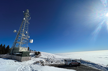 A telecommunications tower and a Strabag service vehicle on a snowy mountain summit above a sea of clouds.