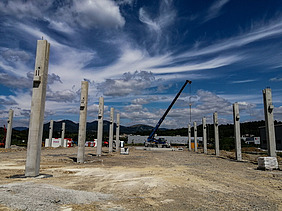 Construction site with multiple upright concrete columns arranged in rows, forming the framework of a large structure. A crane is positioned in the center, lifting materials. The background shows hills, scattered buildings, and a partly cloudy sky with dramatic cloud patterns.