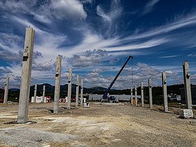 Baustelle mit mehreren aufrecht stehenden Betonsäulen, die in Reihen angeordnet sind und das Gerüst eines großen Bauwerks bilden. In der Mitte steht ein Kran, der Materialien anhebt. Der Hintergrund zeigt Hügel, verstreute Gebäude und einen teilweise bewölkten Himmel mit dramatischen Wolkenmustern.