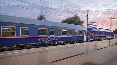 Blauer Nachtzug, der in der Dämmerung über die Bahngleise fährt, mit Bewegungsunschärfe und Überlandleitungen.