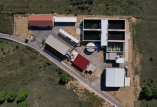 Top-down view of a wastewater treatment facility showing multiple rectangular treatment tanks, circular clarifier, and several buildings with red and white roofs, surrounded by green fields and access roads.