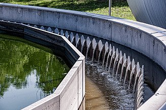 Kreisrundes Klärbecken einer Kläranlage mit Überlaufwehren; klares Wasser fließt über die gezackte Kante, während das angrenzende Becken grünes Wasser enthält - fotografiert auf der Kläranlage Korneuburg.